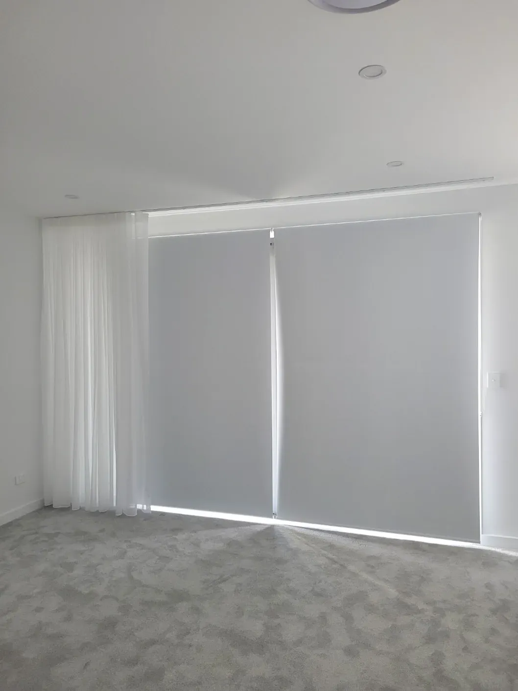 Grey roller blinds paired with a white sheer curtain in a modern carpeted room.