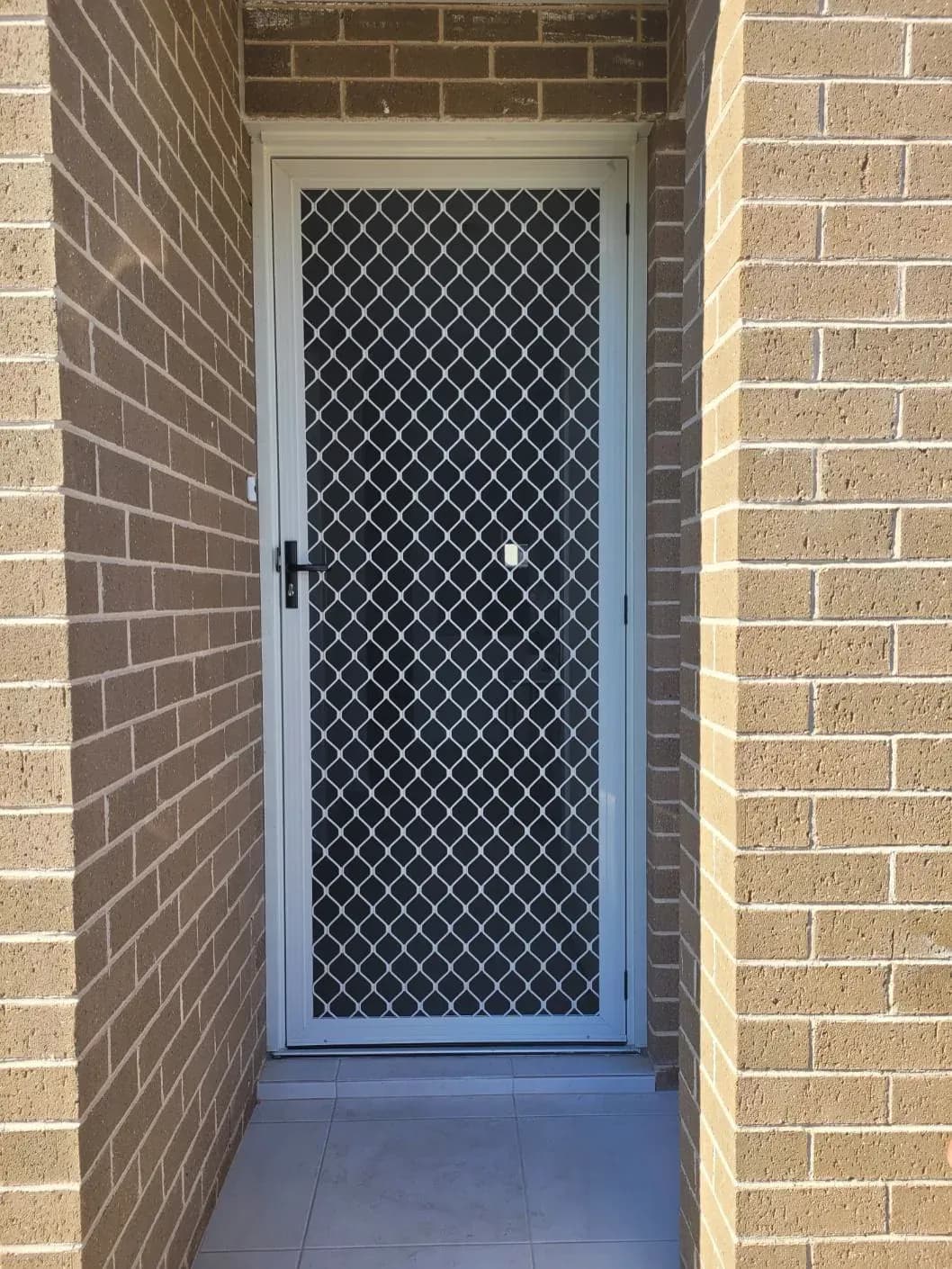 White diamond grille security screen door installed in a brick entryway.