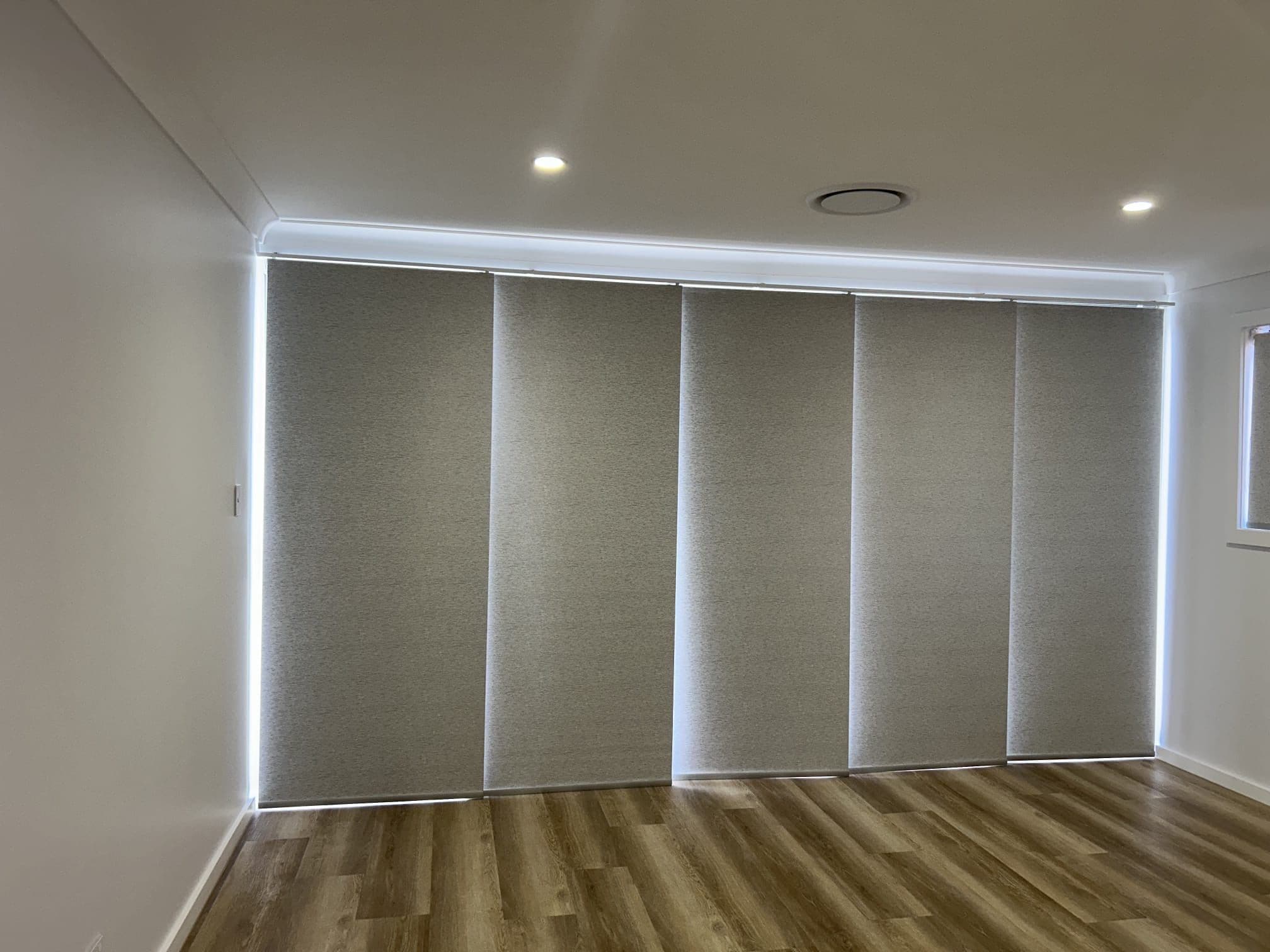 Five grey roller blinds lowered across floor-to-ceiling windows in a room with timber flooring.